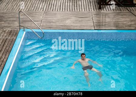 Der junge Mann schwimmt im blauen Swimmingpool im Außenbereich des Hotels. Weißer Kerl aus sportlichem Bauen ist aus der Ferne im Wasser. Tourismus, Urlaubskonzept Stockfoto