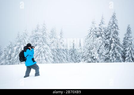 Junger Reisender mit Rucksack fotografiert in einem hohen, schneebedeckten Tannenbaum in hoher Schneeverwehung vor dem Hintergrund von Nebel auf einem frostigen Winter d Stockfoto