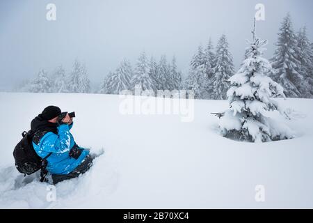 Junger Reisender mit Rucksack fotografiert in einem hohen, schneebedeckten Tannenbaum in hoher Schneeverwehung vor dem Hintergrund von Nebel auf einem frostigen Winter d Stockfoto