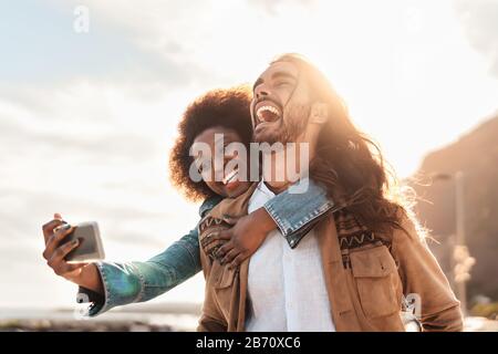 Fröhliches lächelndes Paar, das selfie mit einem mobilen Smartphone im Freien mitnimmt - Junge Trendsleute haben während des Urlaubs Spaß Stockfoto