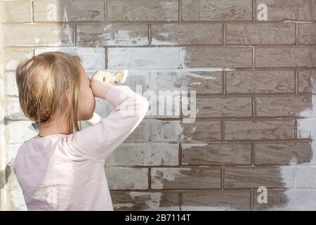 Ein kleines Mädchen wird eine Ziegelwand weiß malen. Wandbemalung, Reparaturen im Haus. Der Maler, der Pflaster malt eine Wand. Abschluss der Fassaden mit Stockfoto