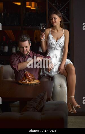 Der Kerl isst eifrig einen Schweineknäuel. Dame, die auf einer Couch sitzt, mit einem Glas Rotwein, auf dunklem Hintergrund in einem Restaurant. Stockfoto