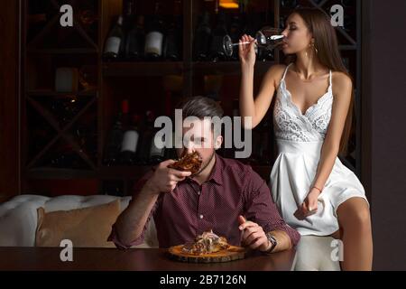 Der Kerl isst eifrig einen Schweineknäuel. Dame, die auf einer Couch sitzt, mit einem Glas Rotwein, auf dunklem Hintergrund in einem Restaurant. Stockfoto
