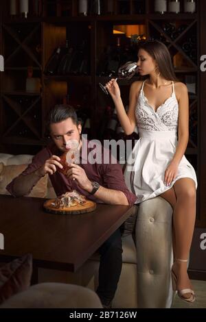 Der Kerl isst eifrig einen Schweineknäuel. Dame, die auf einer Couch sitzt, mit einem Glas Rotwein, auf dunklem Hintergrund in einem Restaurant. Stockfoto