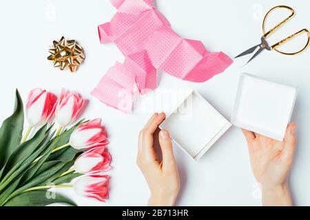 Draufsicht die Hände der Frau, die eine offene Geschenkbox neben zerrissenem Papier, Bogen, Schere und Blumenstrauß auf weißem Tisch hält. Stockfoto