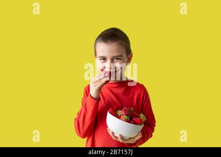 Lächelnder Junge hält eine Erdbeere in Händen auf gelbem Hintergrund, Kind isst rote Beere Stockfoto