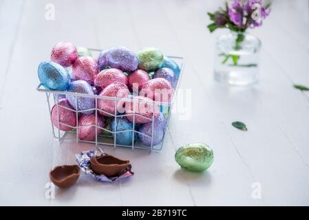 Ein moderner Metallkorb füllte Ostereier aus Schokolade in mehrfarbigem Folienmaterial auf einem weißen Holztisch. Minimalistische festliche Frühlingskomposition. Weich selektiv f Stockfoto