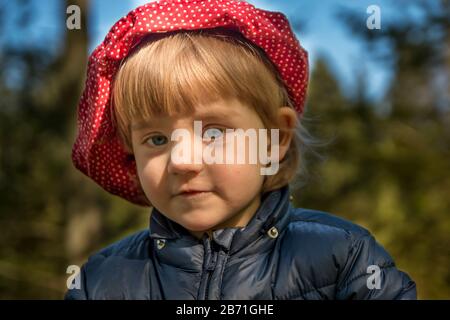 Ein blondes Mädchen von 3 Jahren im Wald, das in die Kamera blickt. Sie trägt einen roten Hut und eine blaue Jacke. Blätter und Bäume im Hintergrund Stockfoto