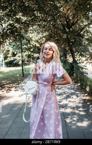 Schöne, stilvolle blonde Frau in polka gepunktet midi-Kleid im städtischen Sommerpark. Sie ist glücklich. Sie trägt eine kleine weiße Tasche und eine schwarze Sonnenbrille. Stockfoto