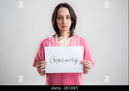 Junger Arzt in rosafarbenem Kleid, der ein weißes Blatt mit Coronavirus-Ankündigung hält und über weißem isoliertem Hintergrund posiert Stockfoto