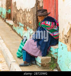 Indigene ältere Tarabuco-Frau am Bürgersteig in der Nähe des sonntagsmarktes mit traditionellem Hut und Textil in Tarabuco, Sucre Department, Bolivien. Stockfoto