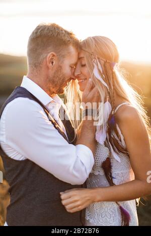 Wunderschönes Paar im Boho-Stil, das sich auf dem sonnigen Sommerfeld umarmte. Gutaussehender Mann in legerer Kleidung und Frau in weißem Kleid und Stockfoto
