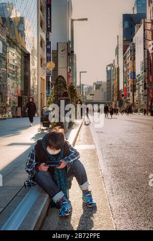 Sehen Sie sich Shin Juku in Tokio an, Wolkenkratzer und Käufer im Hintergrund, als ein kleiner Junge, der eine Maske trägt, auf einem Gutter sitzt, der sein elektronisches Gerät spielt. Stockfoto