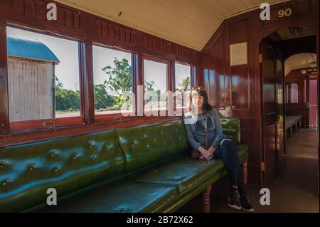 Alleinstehende Touristin sitzt auf einer gepolsterten grünen Sitzbank auf einer alten Holzkutsche, die auf die Züge wartet, die von der Quorn Station in South Australia abfahren. Stockfoto