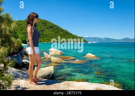 Weiblicher Tourist an einer felsigen Küste auf Fitzroy Island, in Australien, mit Blick auf das schöne türkisfarbene Wasser des Pazifischen Ozeans. Stockfoto