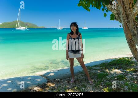 Weiblicher Tourist, der unter einem Baum am Strand steht, der der Kamera mit dem Rücken zum Pazifischen Ozean auf Fitzroy Island, Queensland, zugewandt ist. Stockfoto