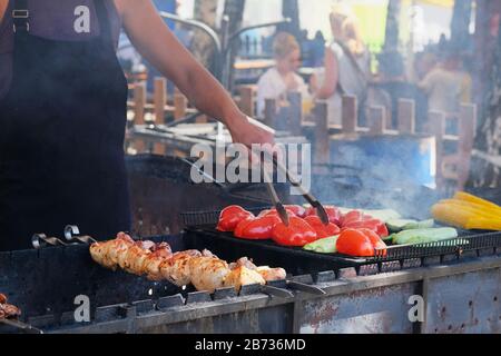 Gegrilltes rotes Gemüse und Fleisch auf der Open-Kitchen-Festveranstaltung. Straßenküche und Kochkonzept für den Außenbereich. Hand des Menschen mit Dongs. Stockfoto