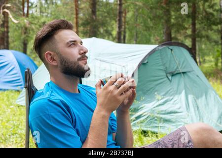 Tourist Kerl hält Tasse auf Zelt und grünen Waldhintergrund. Reise- und Lifestyle-Konzept Stockfoto