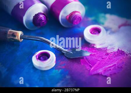 Zwei offene Röhren aus rosa und violettem Ölfarbe liegen neben einem Palettenmesser auf einer in verschiedenen Farben lackierten Palette. Stockfoto