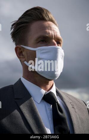 Nahaufnahme eines ernsthaften Geschäftsmannes, der eine chirurgische Gesichtsmaske trägt, die draußen unter grauem Wolkenhimmel steht Stockfoto
