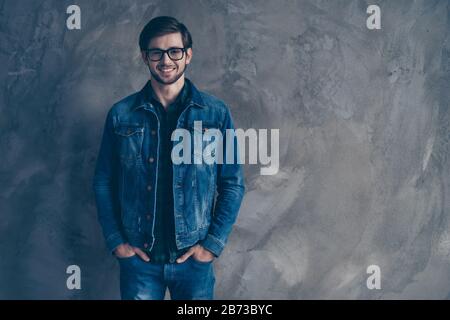 Fröhlicher, trendiger junger, gutaussehender Typ in Brille und mit einem Denimanzug, der auf grauem Hintergrund steht, lächelt und direkt auf die Kamera blickt. Stockfoto