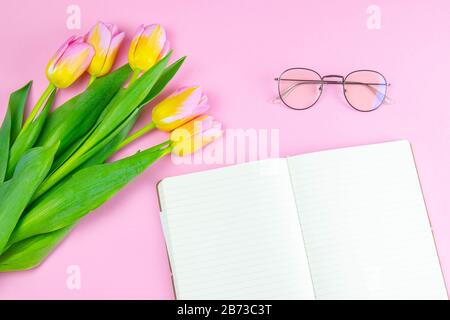 Bunte pinkfarbene und gelbe Tulpe mit offenem Notizbuch und Brille flach lag isoliert auf pinkfarbenem Hintergrund Draufsicht. Frühlingsblume. Womans Day, Mothers Day Stockfoto