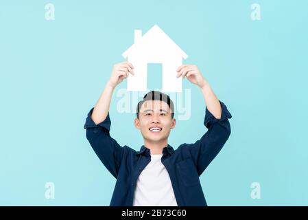 Fröhlicher junger Asiat, der das Schild oben auf hellblauem Hintergrund für Immobilienkonzepte isoliert hält Stockfoto