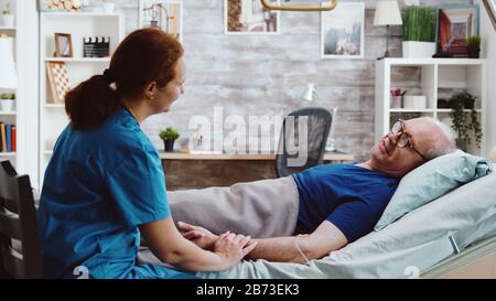 Krankenschwester, die einen Sitz in der Nähe eines alten kranken Mannes nimmt, der im Krankenbett in einem Altenpflegeheim liegt Stockfoto