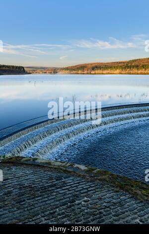 Wasser fließt über abflußkanal von ruhigen malerischen Bäumen gesäumten See, unter tief blauen Himmel - Fewston Reservoir, Washburn Tal, North Yorkshire, England, UK. Stockfoto