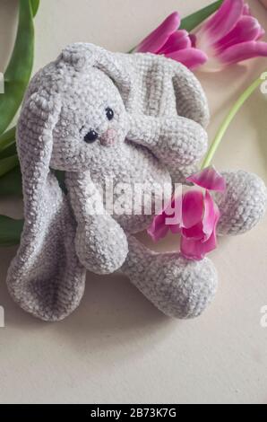 Gestricktes, hellgraues, handgefertigtes Kaninchen mit rosafarbenen Tulpen. Kinderspielzeug. Häkelmuster. Handwerkliche Fertigung. Stockfoto