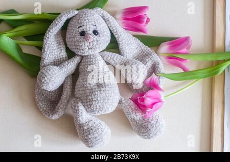 Gestricktes, hellgraues, handgefertigtes Kaninchen mit rosafarbenen Tulpen. Kinderspielzeug. Häkelmuster. Handwerkliche Fertigung. Stockfoto