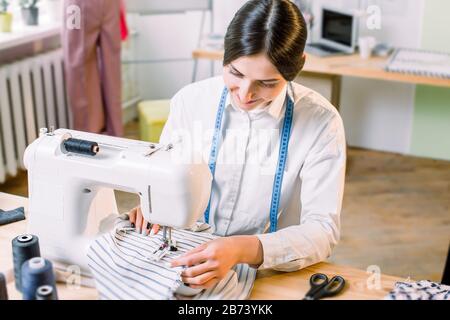 Nahaufnahme des Porträts der jungen Frau, die auf der Nähmaschine sitzt und näht. Mode, Nähkleidung, Hobbynähen als kleines Geschäftskonzept. Stockfoto