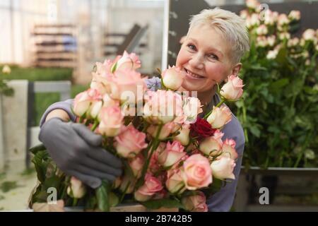 Porträt einer glücklichen reifen Frau mit kurzen Haaren, die an der Kamera lächelt, während sie viele schöne Rosen in ihren Händen hält Stockfoto