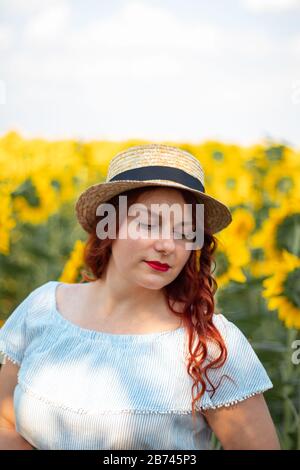 Mädchen in Strohhut und Kleid in Sonnenblumen. Roter Lippenstift, helles Make-up Stockfoto