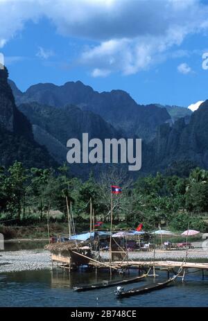 Brücke aus Holz und gewebtem Strohmatt über den Nam Song Fluss bei Vang Vien (Vang Vieng), in der Provinz Vientiane, Laos. Lao-Flaggen und das Reetdach und mehrfarbige Sonnenschirme eines Cafés befinden sich an einer Kieselbank mitten im Fluss. Zwei lange geschweckte Boote sind an die Brücke gebunden. Dahinter befinden sich baumbedeckte, karstbedeckte Berge. Stockfoto