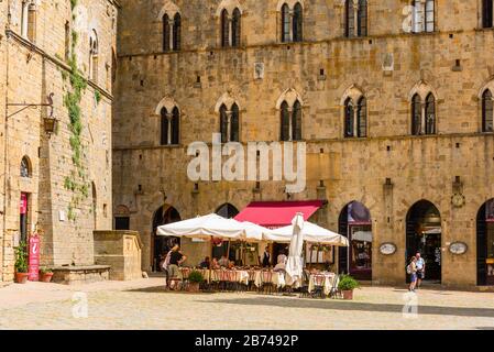 Tische im Schatten vor einem Restaurant auf der Piazza dei priori mit dem Palazzo Pretorio auf der linken Seite in Volterra, Toskana, Italien, an einem Mai-Nachmittag. Stockfoto