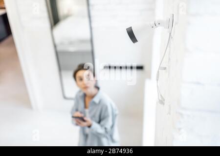 Frau steuert die Überwachung von Heimvideos mit einem Smartphone im Innenbereich, der Ansicht von oben und der ip-Kamera im Vordergrund. Konzept eines drahtlosen Sicherheitssystems für zu Hause Stockfoto