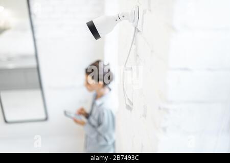 Frau steuert die Überwachung von Heimvideos mit einem Smartphone im Innenbereich, der Ansicht von oben und der ip-Kamera im Vordergrund. Konzept eines drahtlosen Sicherheitssystems für zu Hause Stockfoto