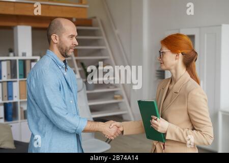 Junge rothaarige Geschäftsfrau in Brillen schüttelt mit Mann die Hände, während sie ein Treffen im Büro haben Stockfoto