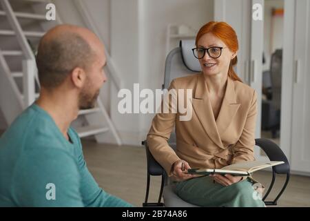 Junge rothaarige Geschäftsfrau in Brillen sitzt auf dem Stuhl und hat ein Interview mit dem Mann im Büro Stockfoto