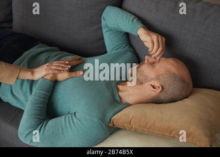Besorgter, reifer Mann, der auf dem Sofa liegt, und Frau, die seine Hand im Wohnzimmer hält Stockfoto