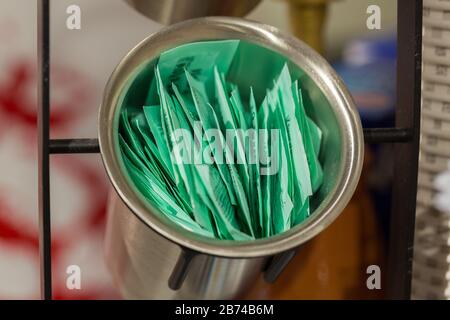 Zuckerbeutel in einer metallischen Schüssel. Viele kleine, grüne Papiersorten, die mit Zucker gefüllt sind. Neutraler Hintergrund, keine Logos. In einem Café. Stockfoto