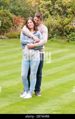 Schöne schwangere junge Frau mit ihrem Lebensgefährten, die sie in ihrem Garten draußen liebkoste Stockfoto