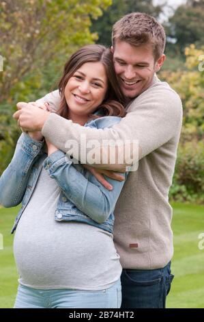 Schöne schwangere junge Frau mit ihrem Lebensgefährten, die sie in ihrem Garten draußen liebkoste Stockfoto