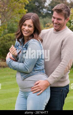 Schöne schwangere junge Frau mit ihrem Lebensgefährten, die sie in ihrem Garten draußen liebkoste Stockfoto