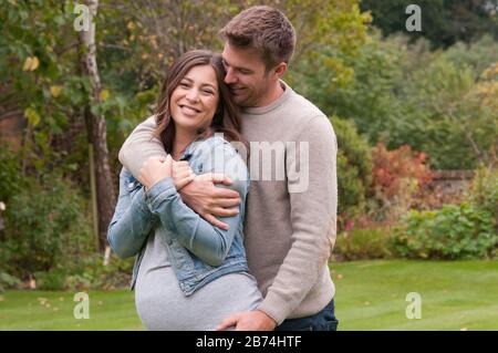 Schöne schwangere junge Frau mit ihrem Lebensgefährten, die sie in ihrem Garten draußen liebkoste Stockfoto