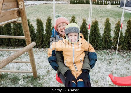 Sohn sitzt auf dem Schoß der Mutter und schwingt draußen im Schnee Stockfoto