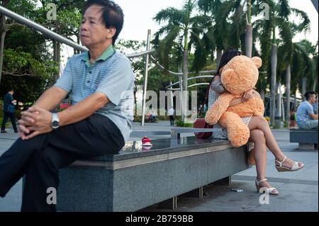 15.03.2019, Singapur, Republik Singapur, Asien - EINE junge Frau sitzt auf einer Bank am Ufer der Marina Bay und umarmt einen Teddybären. [Automatisierte Übersetzung] Stockfoto