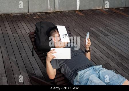 21.07.2018, Singapur, Republik Singapur, Asien - EIN Mann liegt auf einer Sonnenliege in der Marina Bay mit seinem Gesicht bedeckt und schaut auf sein Handy. [Automatisierte Übersetzung] Stockfoto