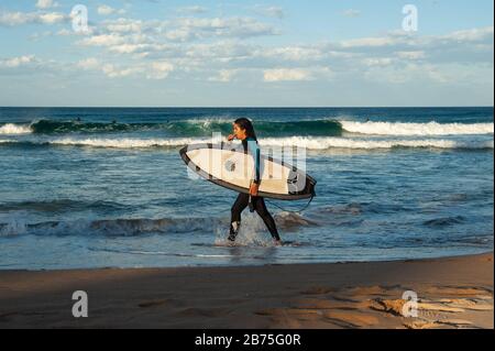 10.05.2018, Sydney, New South Wales, Australien - EINE Frau spaziert am Manly Beach mit ihrem Surfbrett unter dem Arm entlang, während andere Surfer im Hintergrund auf die richtige Welle warten. [Automatisierte Übersetzung] Stockfoto